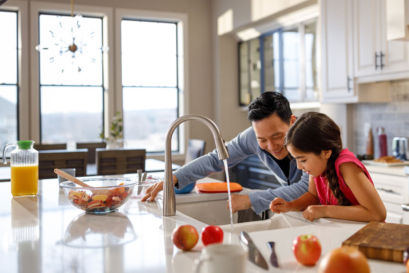 Father and young daughter peer into sink in a light-filled kitchen with faucet water running, both are smiling